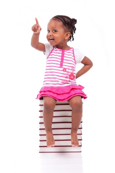 A young girl sitting on books smiling and pointing.