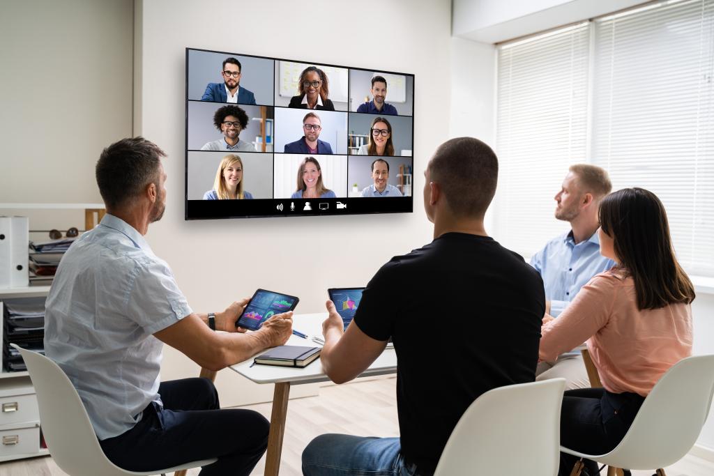 people in a room looking at a big screen with virtual meeting attendees on it